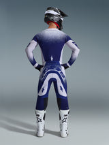 Alpinestars 2026 Supertech Scenz Pants, pantaloni da motocross, grigio chiaro e blu scuro, vista posteriore di un pilota che indossa un completo coordinato con grafiche a sfumatura puntinata, stivali bianchi e casco nero, vestibilità atletica professionale per corse off-road