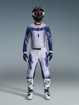 Pilota di motocross che indossa il completo Alpinestars Supertech Scenz 2026, grigio chiaro e blu scuro, composto da maglia e pantaloni con guanti blu coordinati, stivali bianchi e neri e casco nero con occhiali bianchi, outfit coordinato per gare professionali off-road
