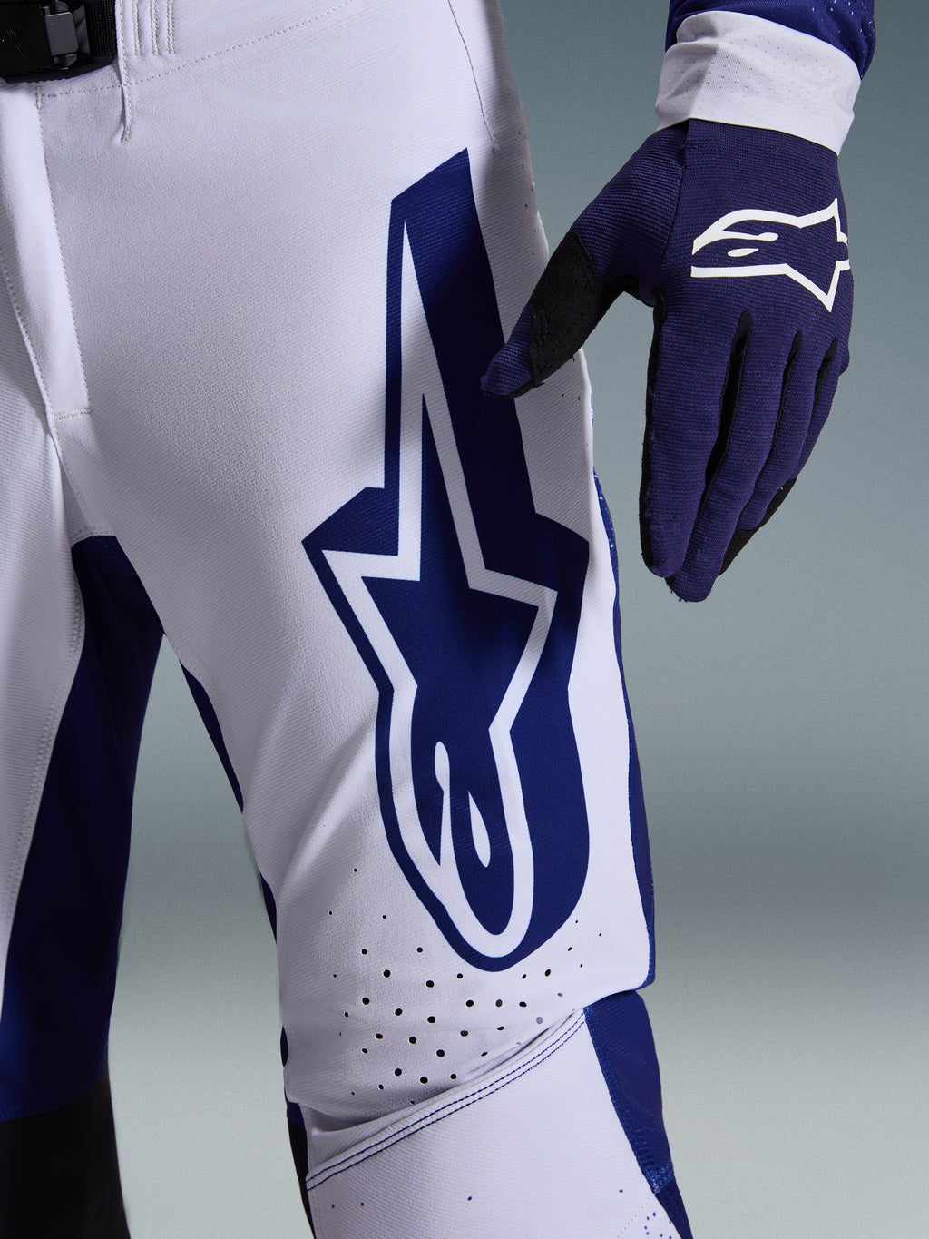 Alpinestars Pantaloni Supertech Scenz 2026, pantaloni da motocross, grigio chiaro e blu scuro, vista ravvicinata della coscia con grande logo Alpinestars blu scuro, zone di ventilazione traforate e tessuto rip-stop elasticizzato, indossati con un guanto da motocross blu scuro coordinato
