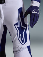 Alpinestars Pantaloni Supertech Scenz 2026, pantaloni da motocross, grigio chiaro e blu scuro, vista ravvicinata della coscia con grande logo Alpinestars blu scuro, zone di ventilazione traforate e tessuto rip-stop elasticizzato, indossati con un guanto da motocross blu scuro coordinato