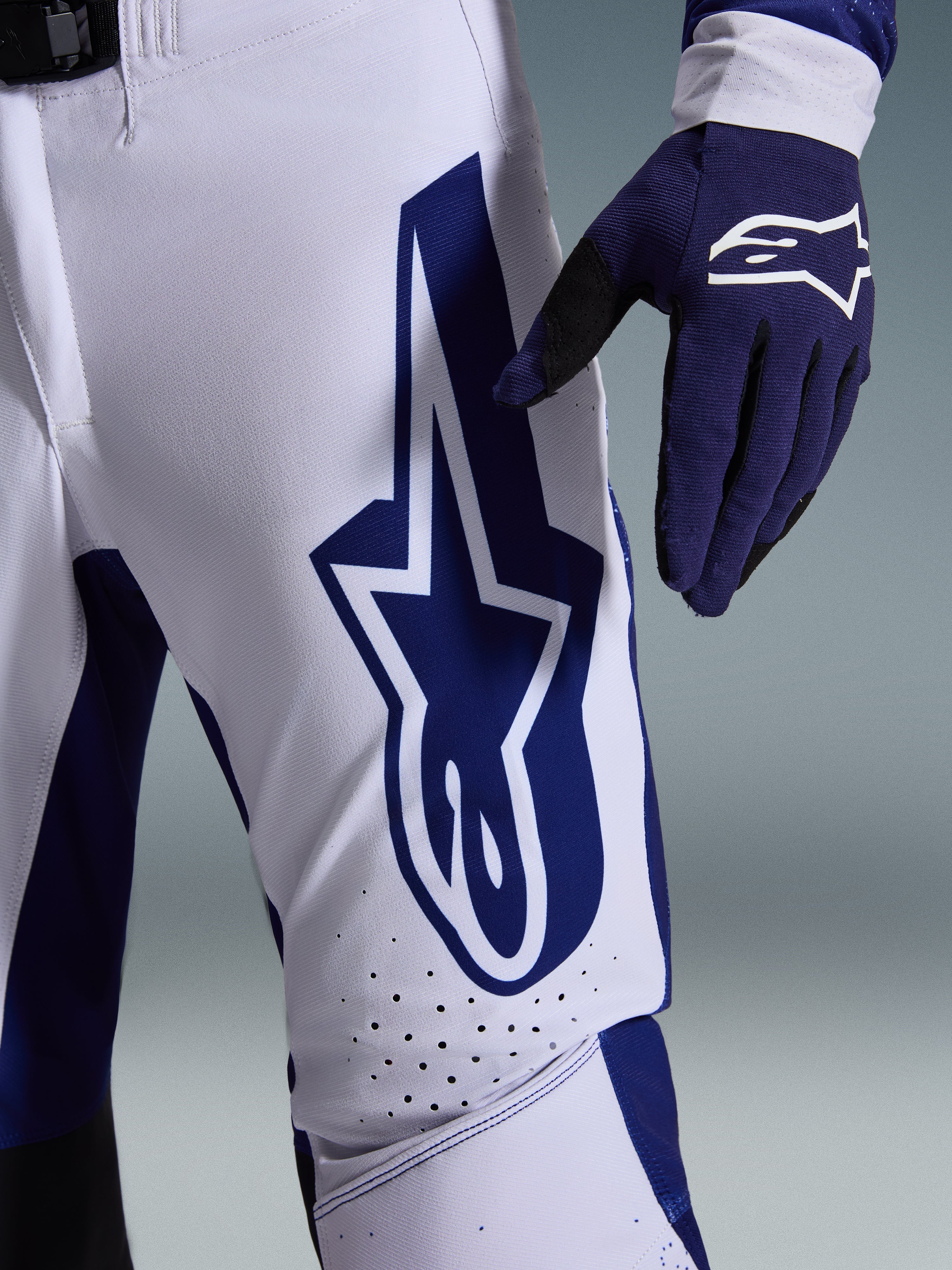 Alpinestars Pantaloni Supertech Scenz 2026, pantaloni da motocross, grigio chiaro e blu scuro, vista ravvicinata della coscia con grande logo Alpinestars blu scuro, zone di ventilazione traforate e tessuto rip-stop elasticizzato, indossati con un guanto da motocross blu scuro coordinato