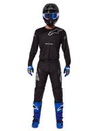 Alpinestars 2025 Racer Graphite Pants, pantaloni da motocross, nero e grigio, indossati da un modello con maglia nera abbinata, stivali blu e viola e occhiali blu, completo da corsa off-road con branding Alpinestars evidente