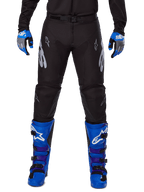 Alpinestars 2025 Racer Graphite Pants, pantaloni da motocross, nero e grigio, indossati con guanti blu e grigi e stivali blu, con branding Astars argento sulle cosce e ginocchia rinforzate, design sportivo per prestazioni off-road