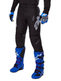Alpinestars Pantaloni Racer Graphite 2025, pantaloni da motocross neri con grafiche bianche e grigie, indossati con stivali da motocross blu acceso e guanti blu coordinati, tessuto tecnico resistente per il fuoristrada