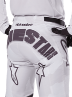 Vista posteriore dei pantaloni Alpinestars Racer Hollow 2025, pantaloni da motocross, bianco e mimetico chiaro, con grande marchio Alpinestars nero sulla seduta, indossati con maglia coordinata e guanti neri