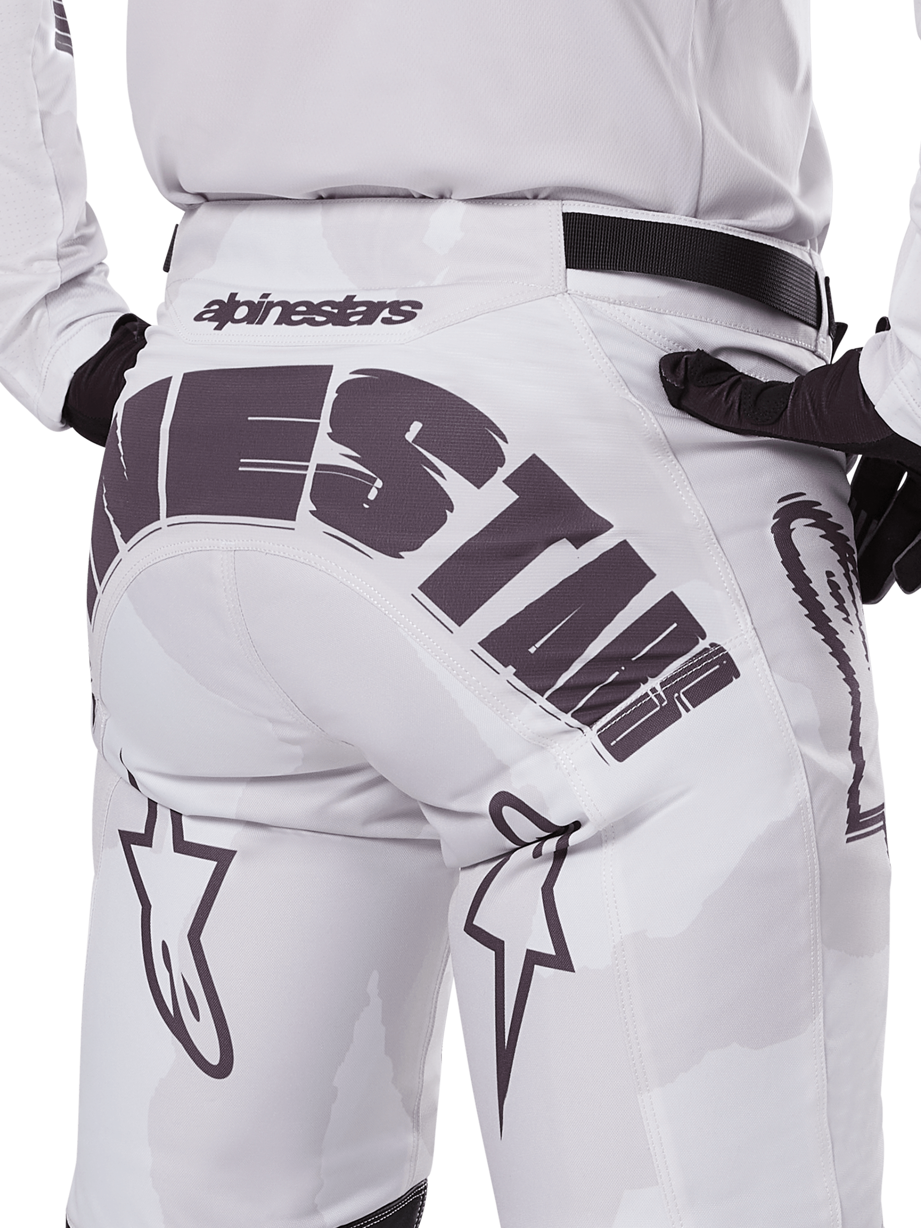 Vista posteriore dei pantaloni Alpinestars Racer Hollow 2025, pantaloni da motocross, bianco e mimetico chiaro, con grande marchio Alpinestars nero sulla seduta, indossati con maglia coordinata e guanti neri