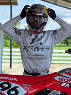 Pilota di motocross con abbigliamento Alpinestars Racer Hollow 2025, maglia camouflage chiaro con grande logo ASTARS, mentre regola un casco Red Bull con maschera trasparente, in piedi accanto a una moto da cross Honda rossa in un contesto off-road.
