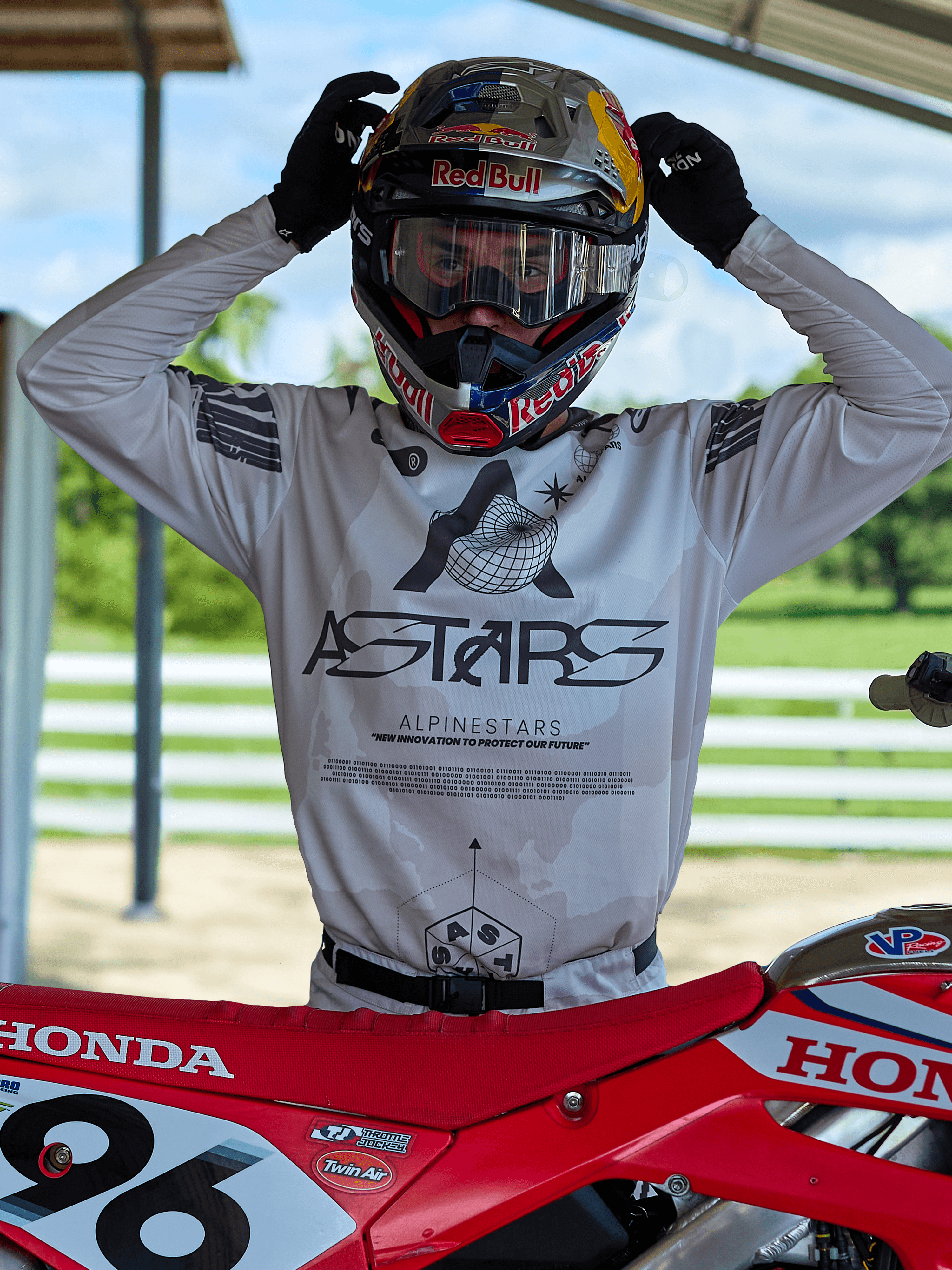 Pilota di motocross con abbigliamento Alpinestars Racer Hollow 2025, maglia camouflage chiaro con grande logo ASTARS, mentre regola un casco Red Bull con maschera trasparente, in piedi accanto a una moto da cross Honda rossa in un contesto off-road.
