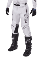 Pantaloni Alpinestars Racer Hollow 2025, pantaloni da motocross, camo bianco e grigio, con grandi loghi a stella profilati in nero e pannelli interni alle ginocchia in pelle nera, indossati con stivali e guanti da motocross neri, design resistente per corse fuoristrada.