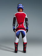 Alpinestars 2026 Techstar Knif Pants, pantaloni da motocross, nero bianco rosso e blu, vista posteriore di un pilota in equipaggiamento completo, maglia coordinata, casco blu e stivali neri e rossi, con vistoso marchio Alpinestars sulla seduta.