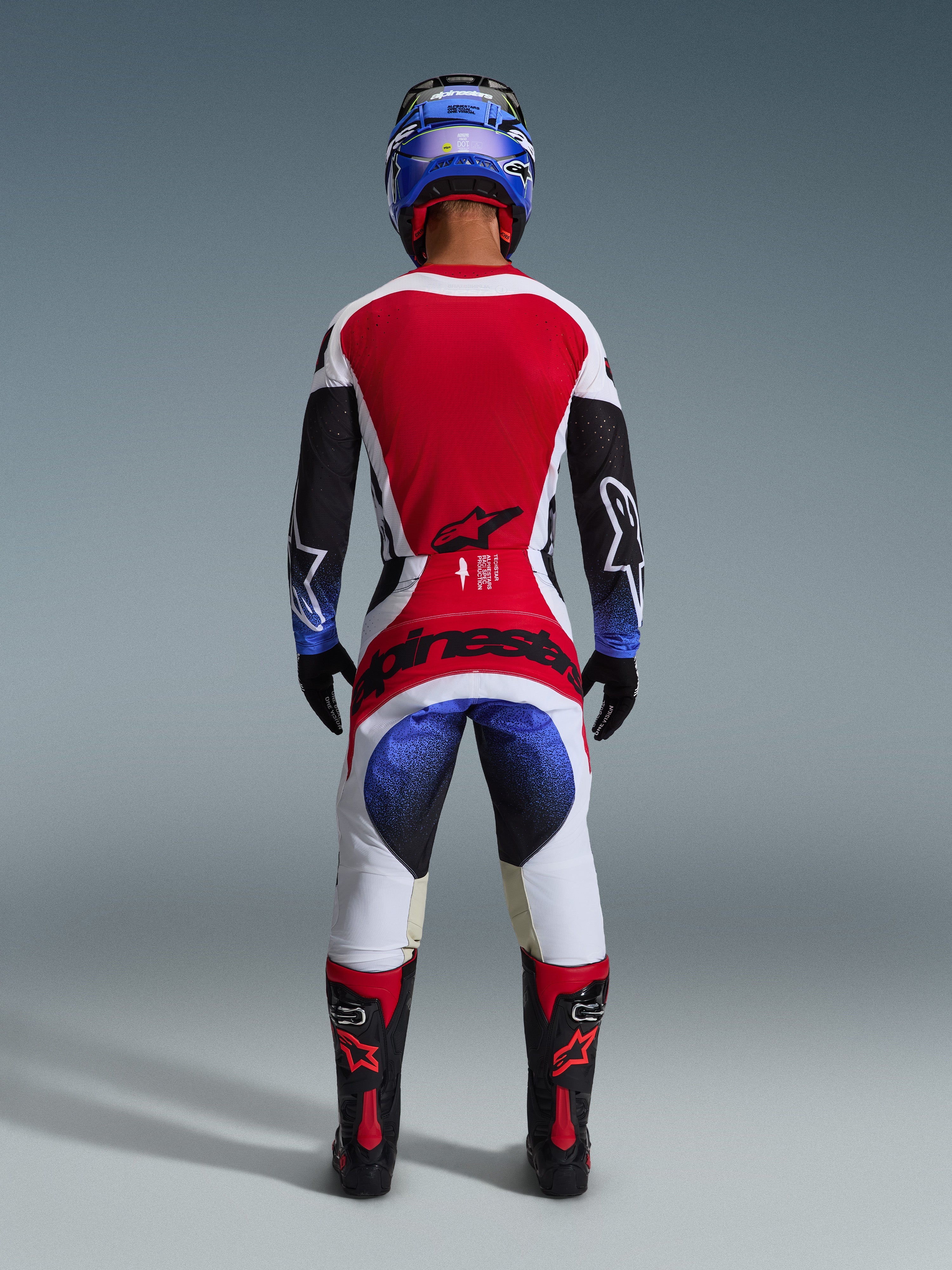 Alpinestars 2026 Techstar Knif Pants, pantaloni da motocross, nero bianco rosso e blu, vista posteriore di un pilota in equipaggiamento completo, maglia coordinata, casco blu e stivali neri e rossi, con vistoso marchio Alpinestars sulla seduta.
