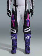 Pilota di motocross che indossa il completo Alpinestars Techstar Knif 2026, pantaloni bianchi e neri con loghi rosa e blu, stivali viola con marchio bianco, guanti neri, vestibilità race atletica e ventilazione tagliata al laser per gare fuoristrada