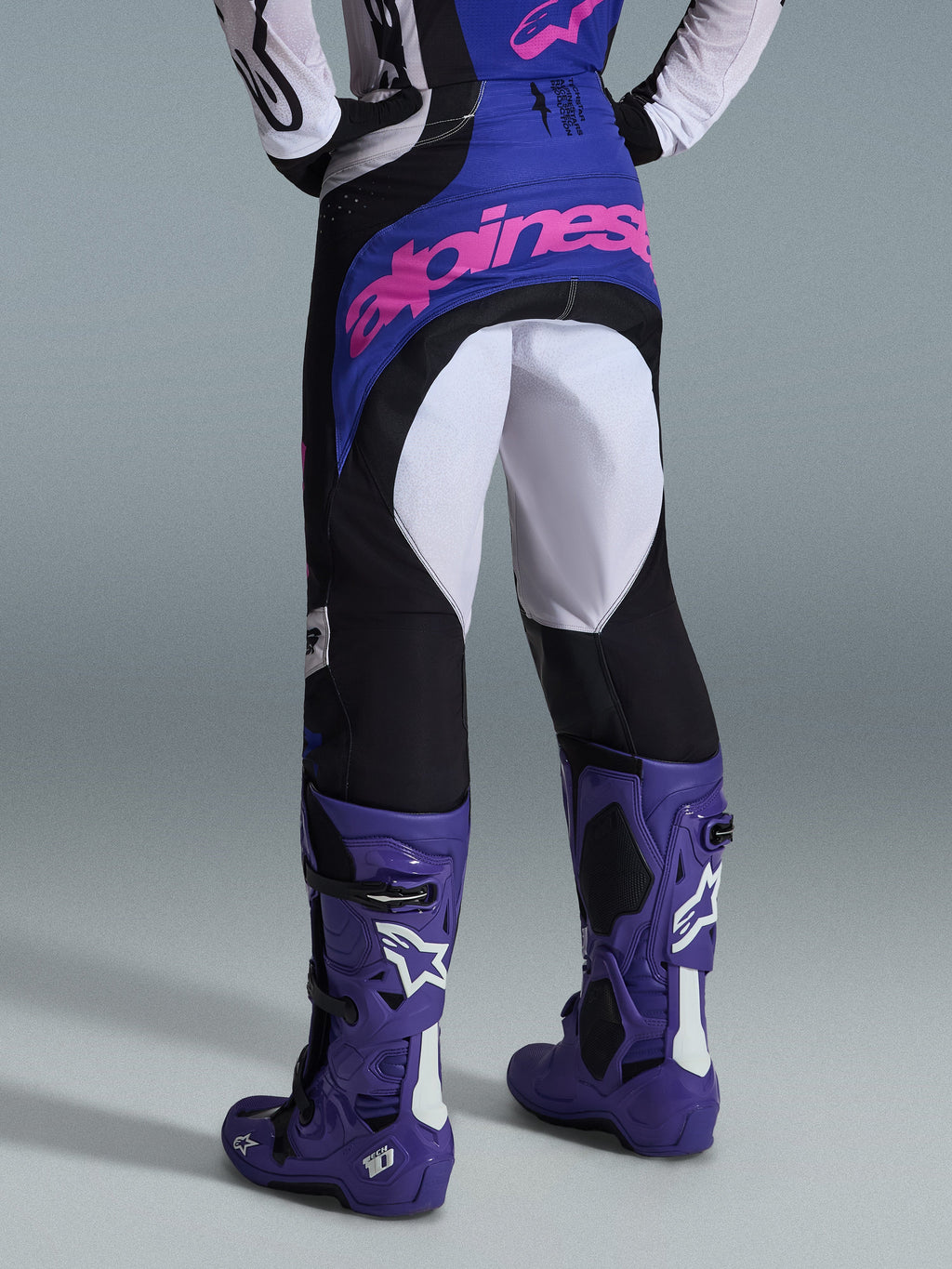 Alpinestars Techstar Knif 2026, pantaloni da motocross, nero, grigio chiaro e viola, vista posteriore con logo rosa sulla seduta, indossati con maglia coordinata e stivali da motocross viola, completo professionale da gara off-road