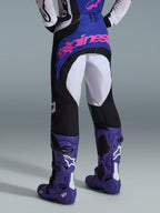 Alpinestars Techstar Knif 2026, pantaloni da motocross, nero, grigio chiaro e viola, vista posteriore con logo rosa sulla seduta, indossati con maglia coordinata e stivali da motocross viola, completo professionale da gara off-road