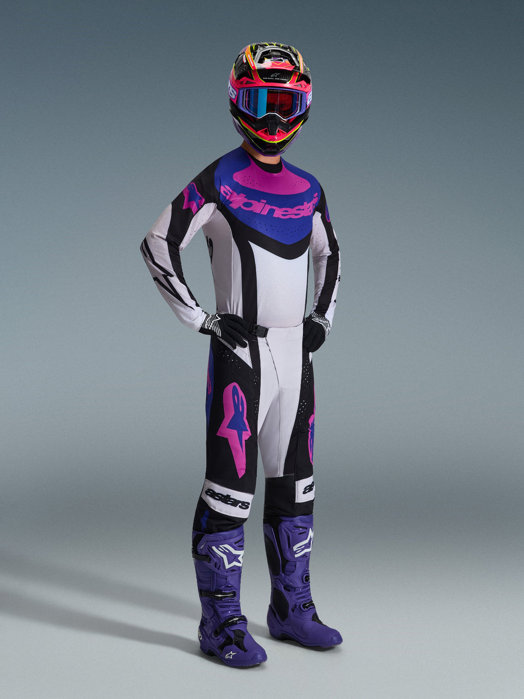 Pilota di motocross con il set completo Alpinestars Techstar Knif 2026, maglia bianca con dettagli neri, blu e rosa, pantaloni coordinati con loghi Astars rosa, casco multicolore con occhiali, stivali da gara viola, abbigliamento tecnico ad alte prestazioni per motocross