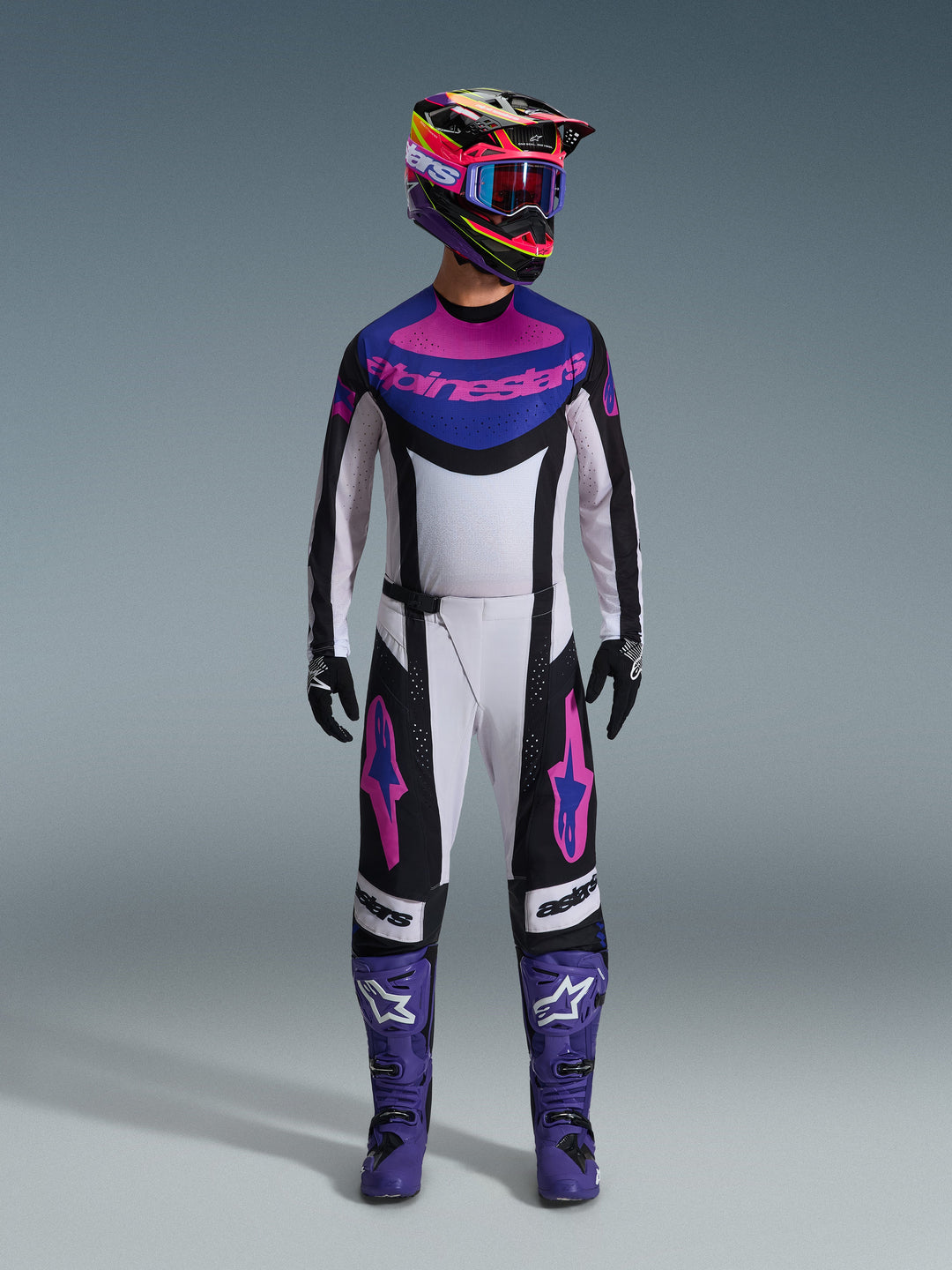 Alpinestars 2026 Techstar Knif Gear Set, completo da motocross, bianco, nero, viola e rosa, indossato con casco multicolore, maschera blu, stivali viola e guanti neri, kit coordinato ad alte prestazioni per gare fuoristrada