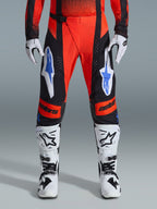 Alpinestars 2026 Techstar Knif Pants, pantaloni da motocross, arancione, nero e blu UCLA, indossati con maglia coordinata, stivali da motocross bianchi e neri e guanti, con logo evidente su cosce e ginocchia, vestibilità sportiva da gara per corse off-road