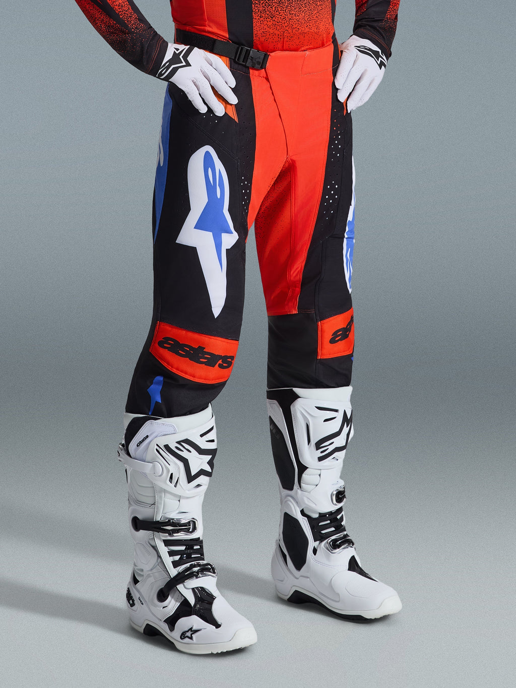 Alpinestars 2026 Techstar Knif Pants, pantaloni da motocross, arancione, nero e blu UCLA, indossati con guanti bianchi e stivali da motocross bianchi e neri, con loghi evidenti sulle cosce e pannelli sulle ginocchia arancioni con marchio astars.