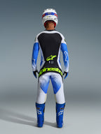 Pilota di motocross visto da dietro con equipaggiamento da gara completo, pantaloni Techstar Knif 2026 in UCLA Blue, nero e giallo fluo, maglia coordinata a maniche lunghe con pannelli bianchi e neri, stivali da fuoristrada blu e casco bianco, con evidente marchio Alpinestars giallo fluo sulla seduta.