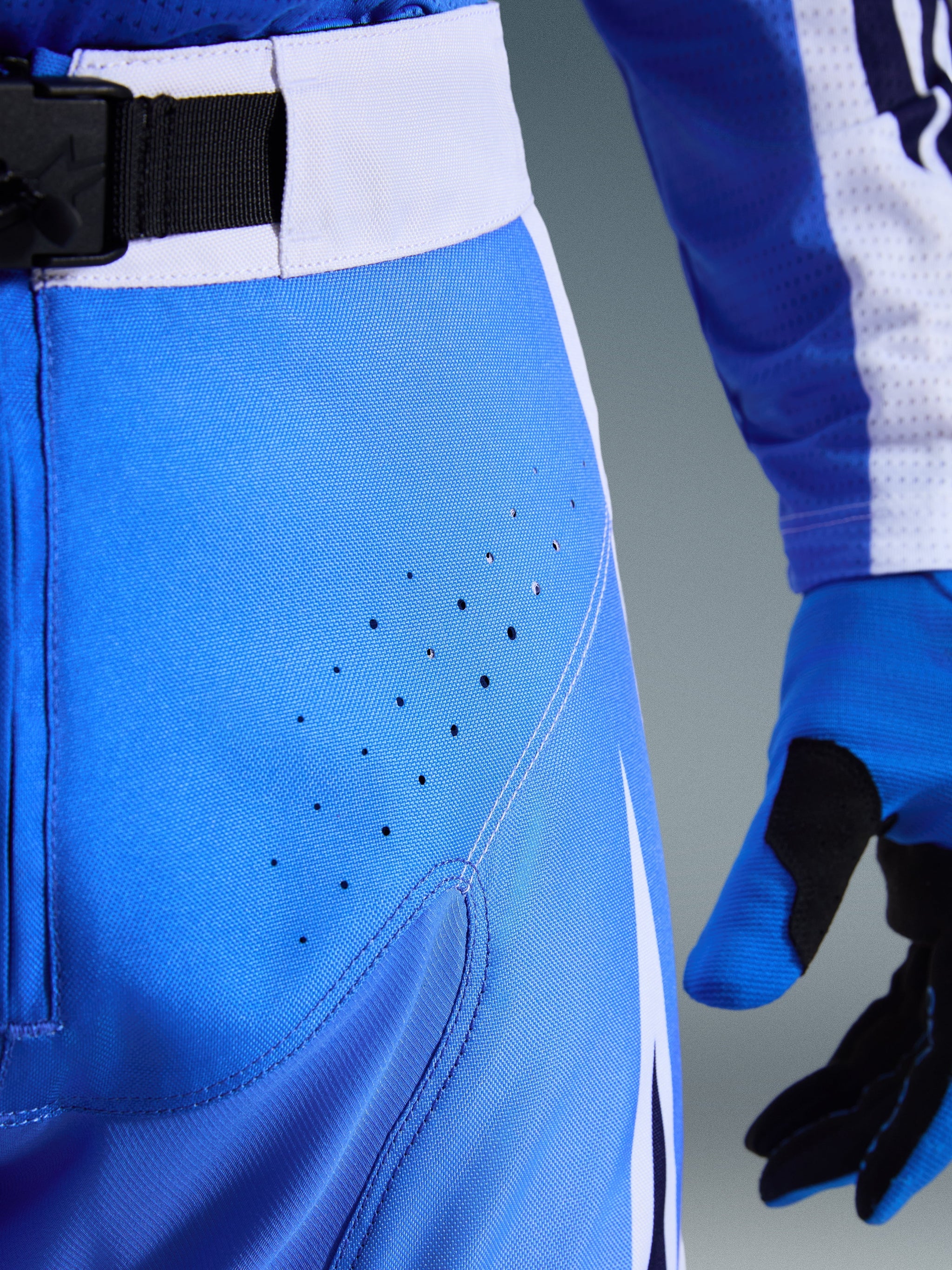 Alpinestars 2026 Techstar Knif Pants, blu Ucla e bianco, primo piano dell'area dell'anca con perforazioni tagliate al laser per la ventilazione, chiusura in vita con fibbia magnetica SX2 e guanti da motocross coordinati