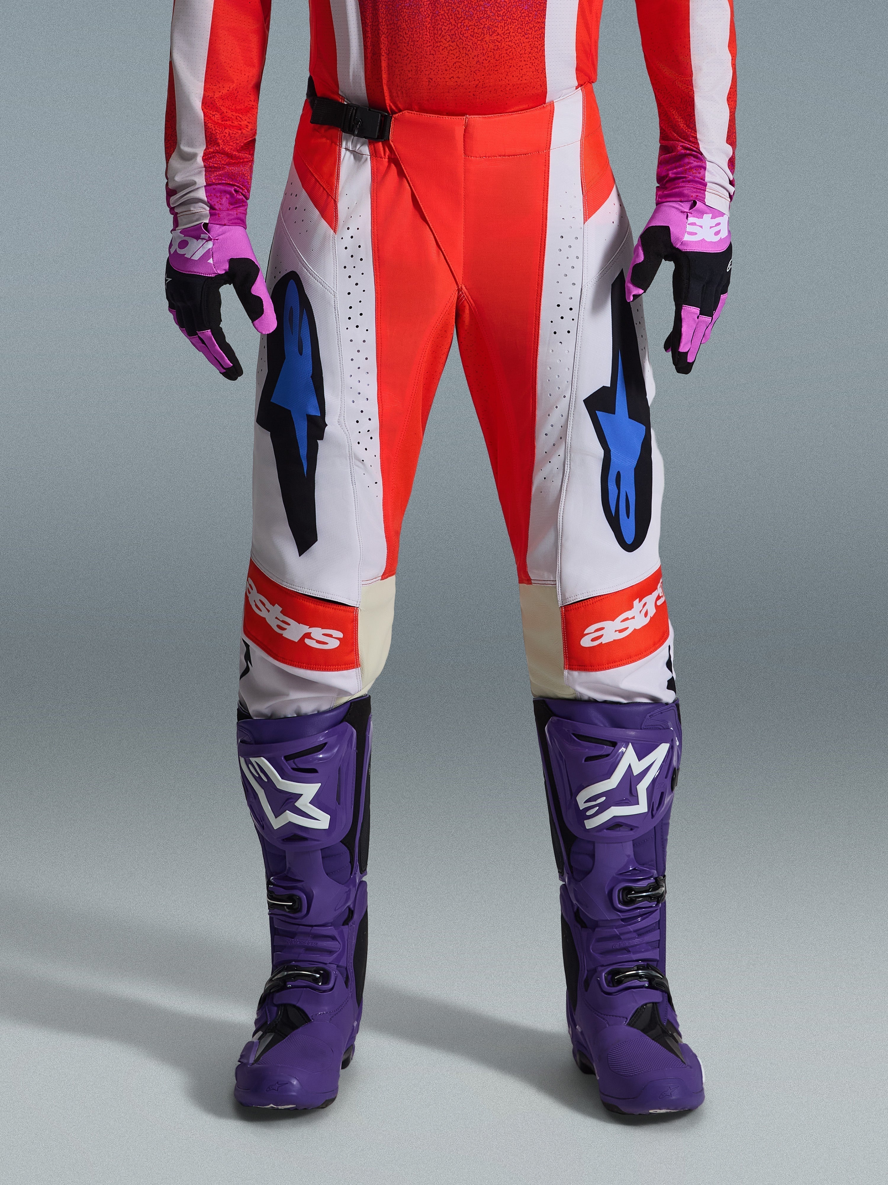 Alpinestars Pantaloni Techstar Knif 2026, pantaloni da motocross in arancione fluo e grigio chiaro con accenti neri, mostrati con stivali e guanti da motocross viola, caratterizzati da loghi a stella blu e costruzione multi-pannello per prestazioni off-road