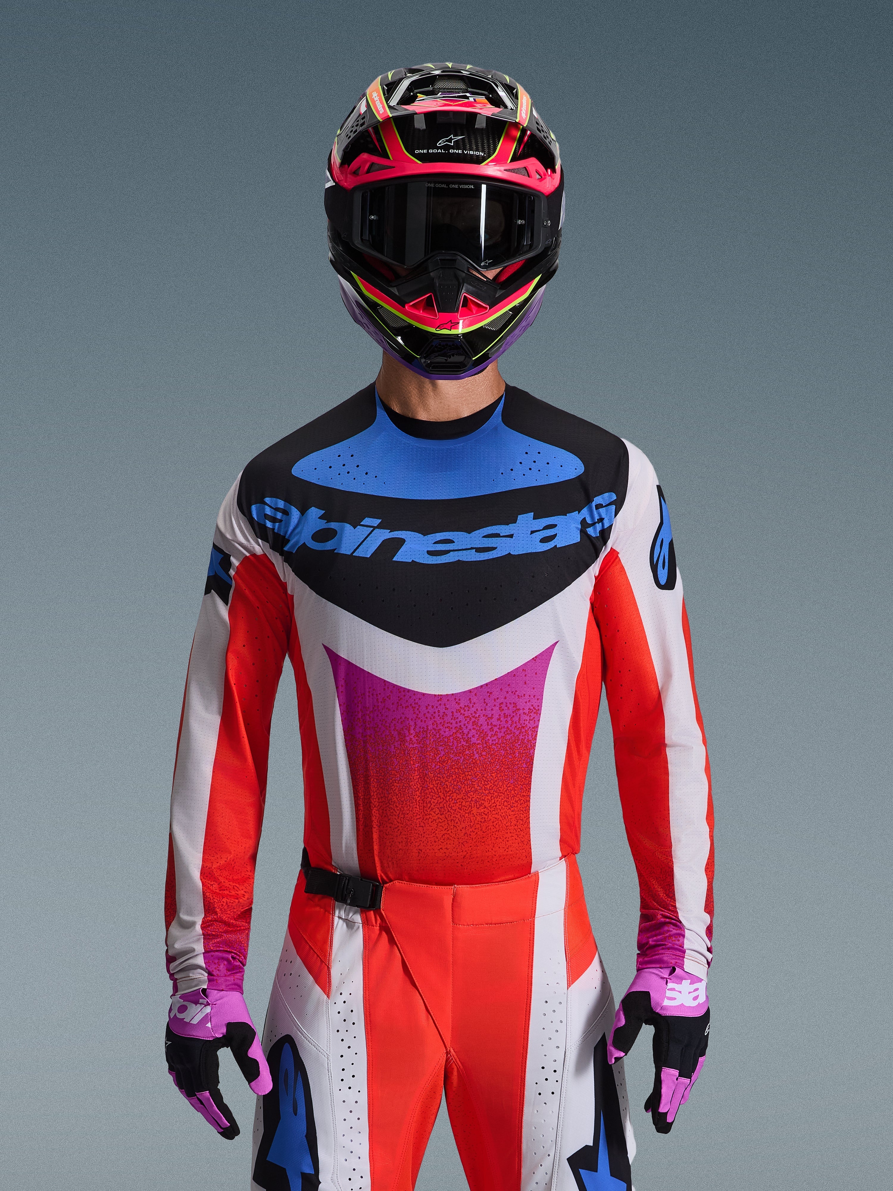 Pilota di motocross che indossa la maglia Alpinestars Techstar Knif 2026 nella colorazione Light Gray Orange Fluo Black, caratterizzata da un vistoso logo blu e vivaci maniche arancioni, abbinata a un casco rosa e nero e guanti viola, vestibilità atletica da gara per competizioni off-road ad alte prestazioni