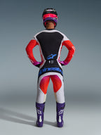 Pilota di motocross visto da dietro con pantaloni Techstar Knif 2026 e maglia abbinata, design grigio chiaro e nero con accenti arancione fluo e viola, branding Alpinestars blu sulla seduta, stivali e casco viola, set completo professionale per gare off-road