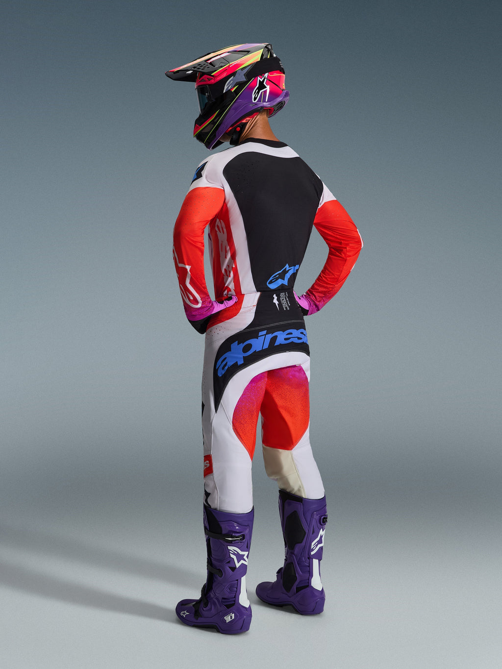 Pilota di motocross visto da dietro con abbigliamento completo da gara, pantaloni Alpinestars 2026 Techstar Knif in grigio chiaro e arancione fluo con loghi blu, maglia coordinata con maniche arancioni, stivali Tech 10 viola e casco da corsa colorato, outfit off-road professionale coordinato