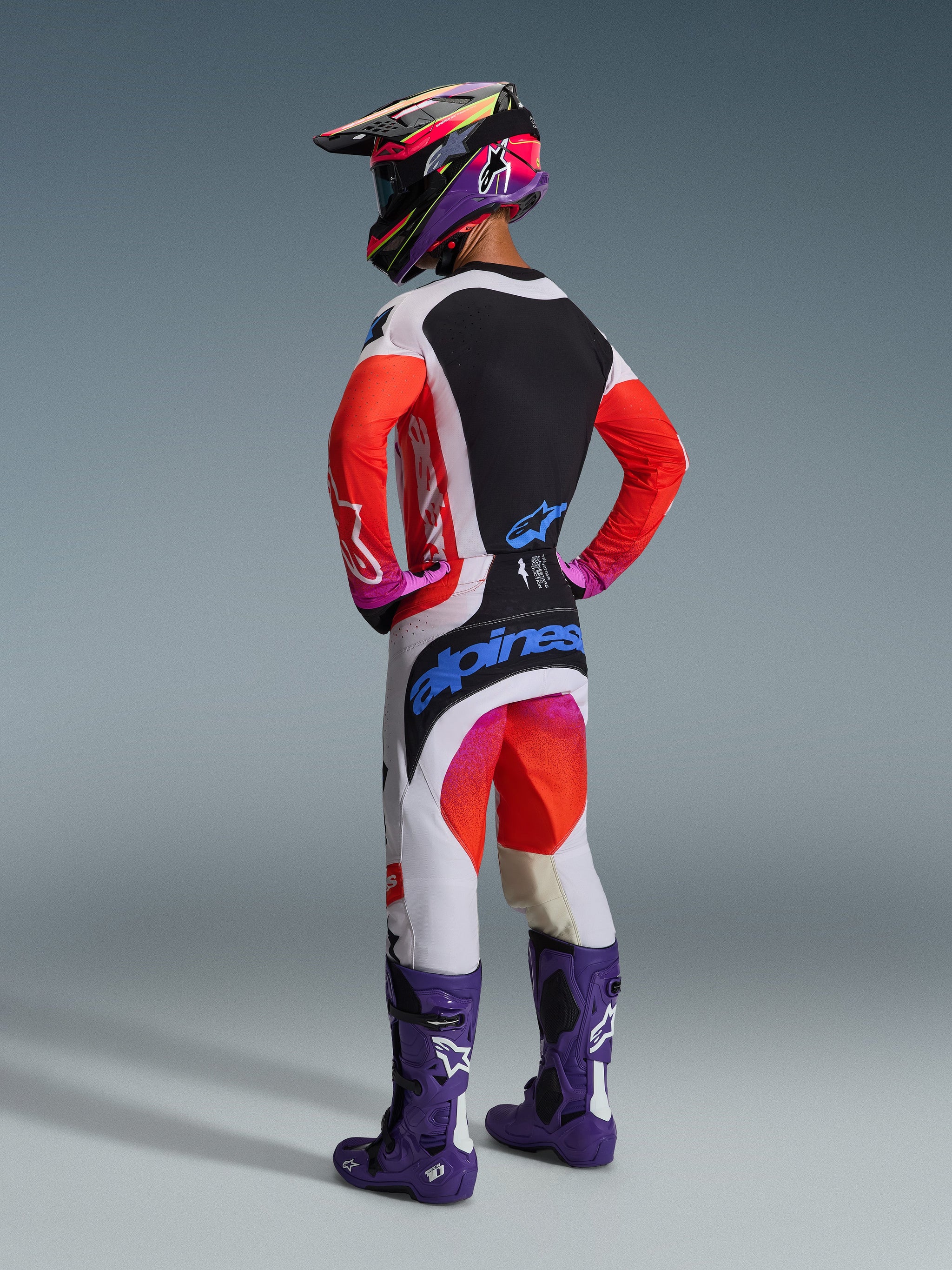 Vista posteriore di un pilota di motocross che indossa la maglia Alpinestars 2026 Techstar Knif, grigio chiaro e nero con maniche arancione fluorescente, abbinata a pantaloni da gara coordinati, stivali viola e casco multicolore, set professionale completo da off-road.