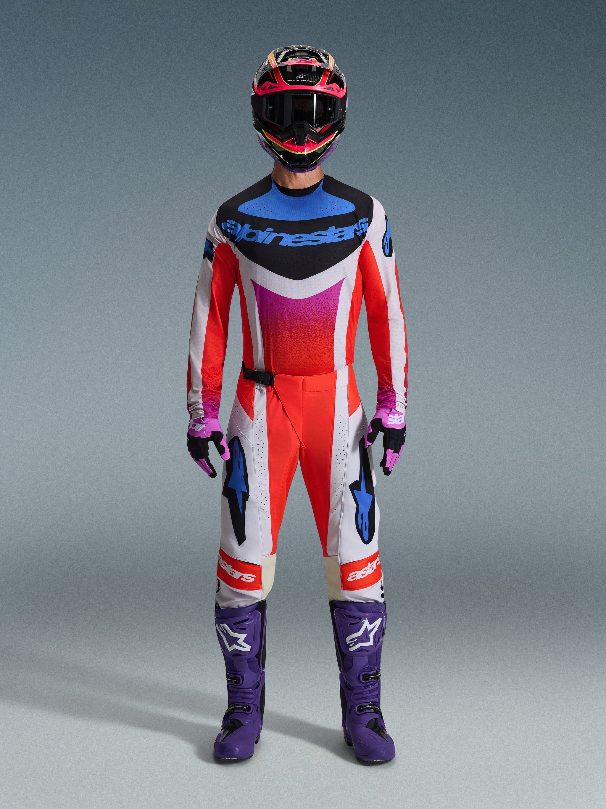 Pilota di motocross con abbigliamento da gara completo, maglia Alpinestars 2026 Techstar Knif e pantaloni coordinati in bianco e arancione con accenti blu e viola, stivali da corsa viola con logo bianco e casco multicolore, outfit coordinato per competizioni off-road