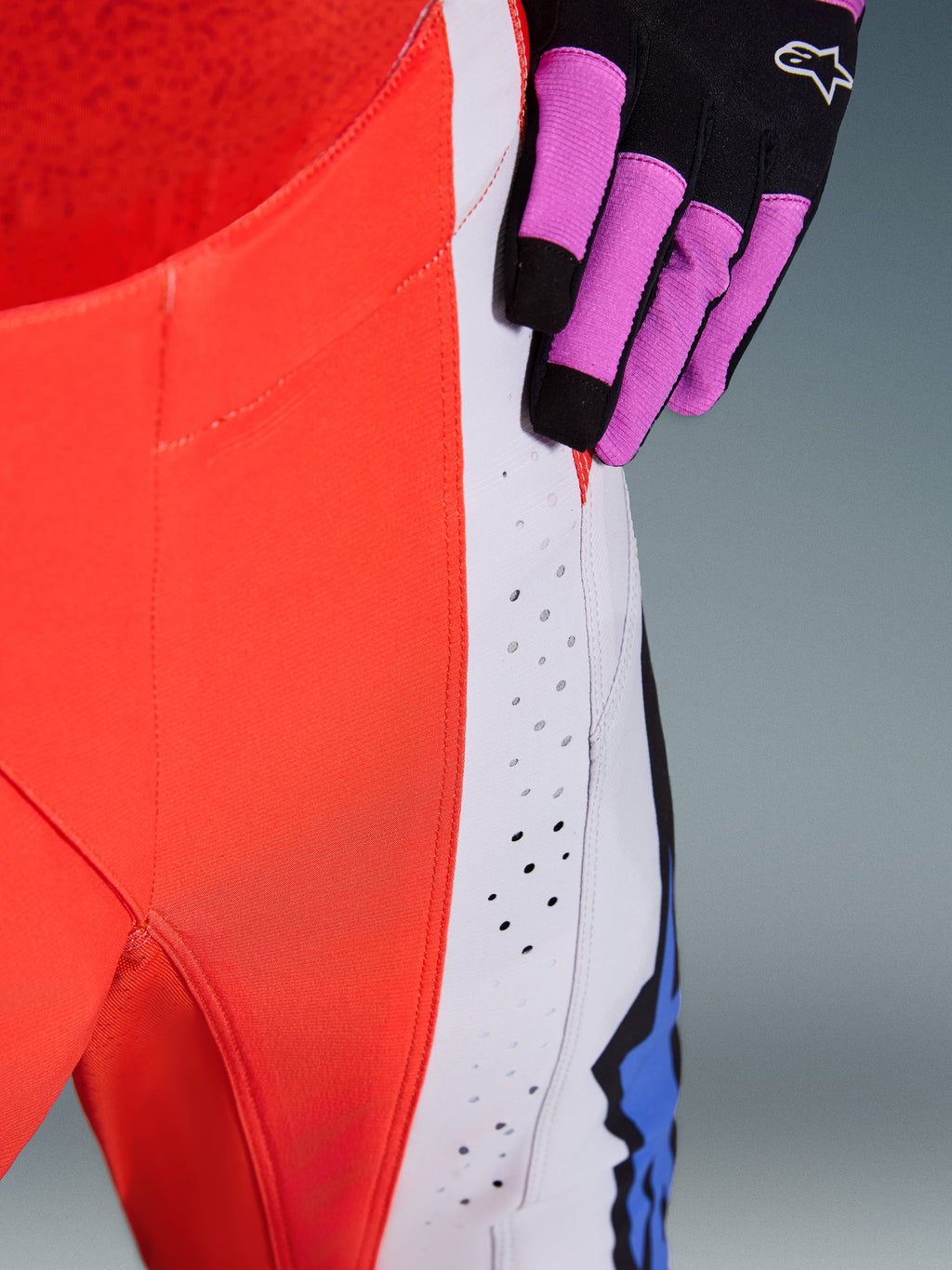 Alpinestars 2026 Techstar Knif Pants, pantaloni da motocross, arancione fluo e grigio chiaro con accenti blu, primo piano dei fori di ventilazione tagliati al laser sulla coscia, indossati con un guanto da motocross nero e viola
