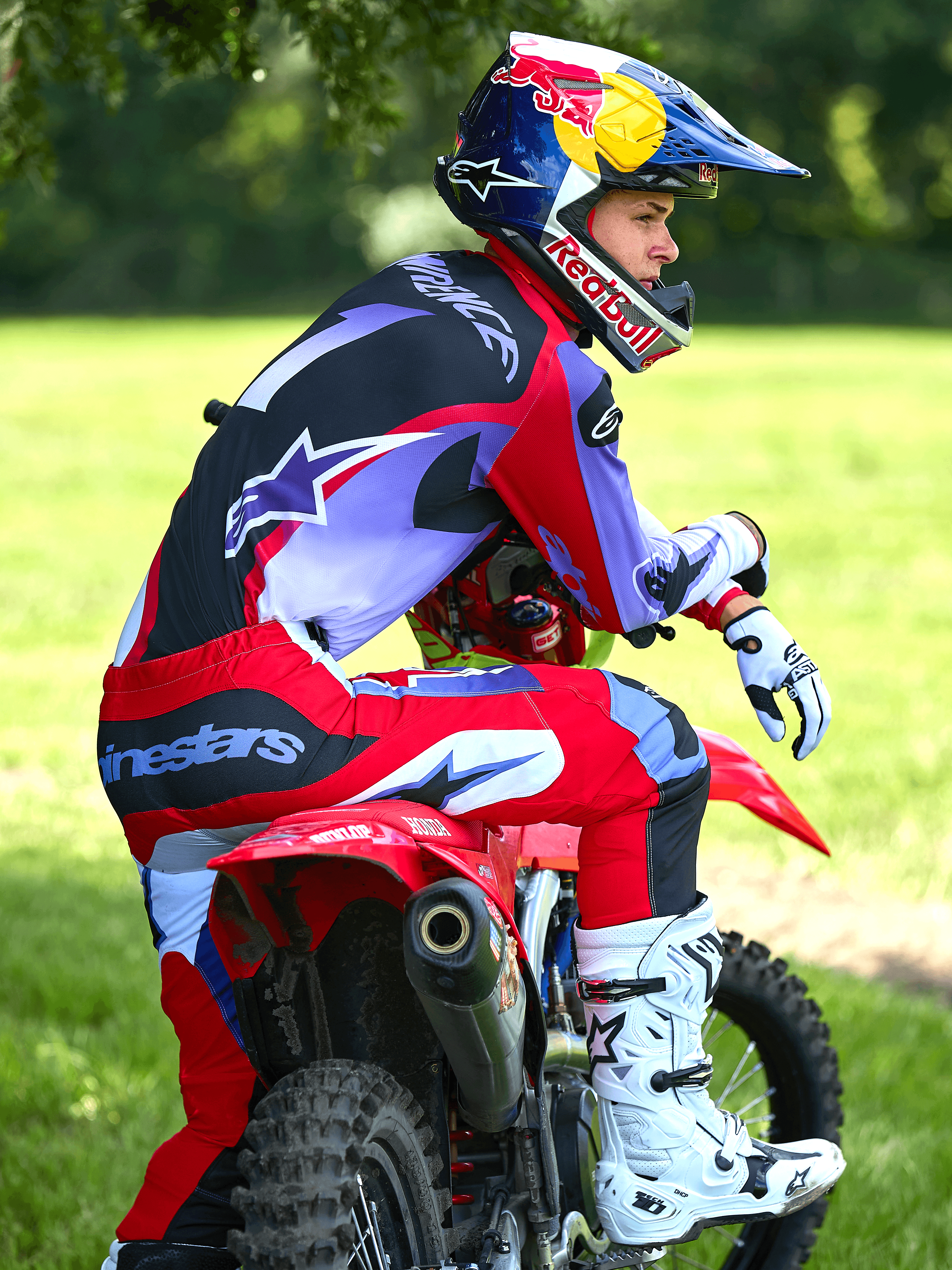 Pilota di motocross seduto su una moto da cross rossa, indossa i pantaloni Alpinestars 2025 Fluid Wurx in viola e rosso brillante, maglia coordinata, stivali Tech 10 bianchi e neri, guanti bianchi e casco Red Bull, completo da gara off-road in un ambiente all'aperto