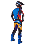 Pilota di motocross visto da dietro con pantaloni Alpinestars 2025 Fluid Wurx, design multicolore con dettagli blu, arancioni, gialli e viola, maglia abbinata, stivali blu acceso e casco nero, equipaggiamento completo per fuoristrada