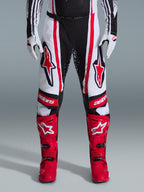 Alpinestars pantaloni Techstar Nomur 2026, pantaloni da motocross, nero, bianco e rosso, indossati con stivali rossi e guanti bianchi, con audaci loghi rossi sulle cosce e marchio 'astars' sulle ginocchia, design racing multi-pannello per prestazioni off-road