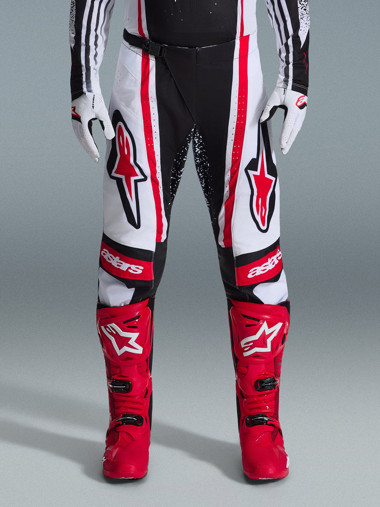 Alpinestars pantaloni Techstar Nomur 2026, pantaloni da motocross, nero, bianco e rosso, indossati con stivali rossi e guanti bianchi, con audaci loghi rossi sulle cosce e marchio 'astars' sulle ginocchia, design racing multi-pannello per prestazioni off-road