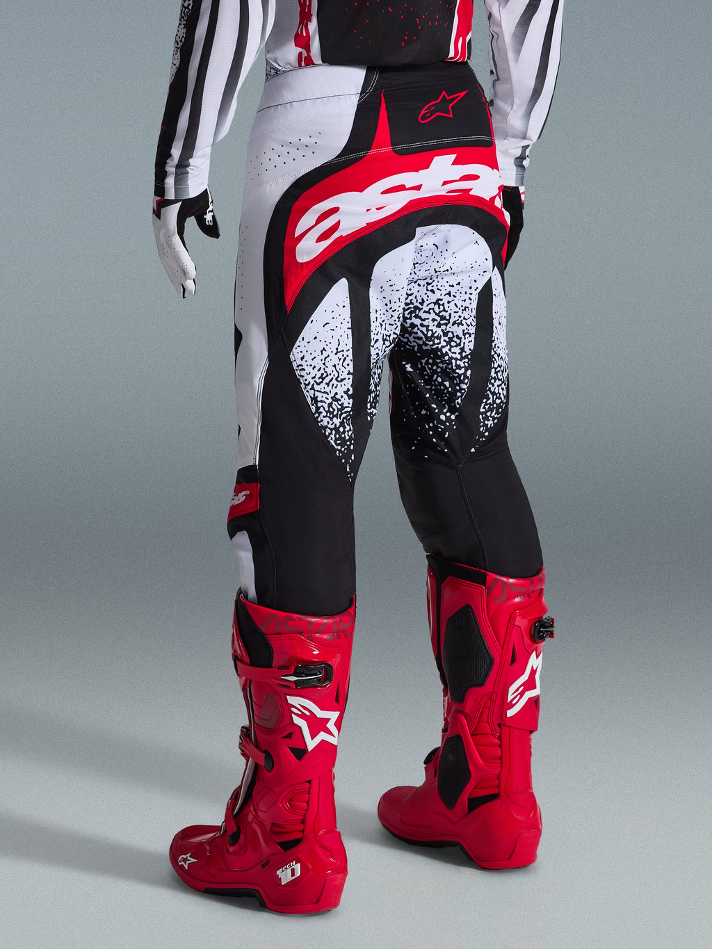 Pantaloni Alpinestars Techstar Nomur 2026, pantaloni da motocross, nero, bianco e rosso, vista posteriore su un pilota con stivali rossi coordinati, caratterizzati da un grande logo 'astars' rosso e bianco sulla seduta e grafiche punteggiate, equipaggiamento professionale per competizioni fuoristrada
