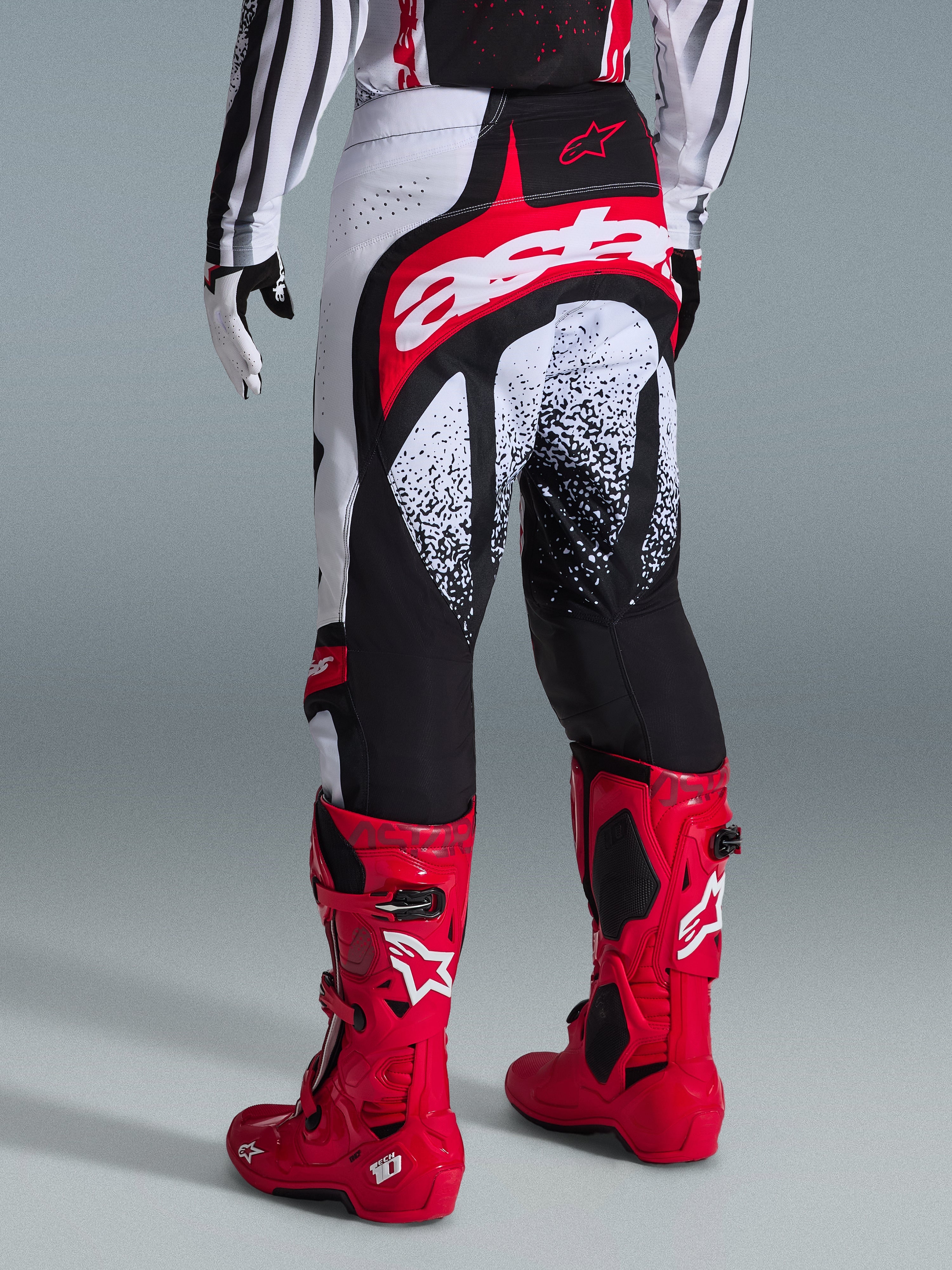 Pantaloni Alpinestars Techstar Nomur 2026, pantaloni da motocross, nero, bianco e rosso, vista posteriore su un pilota con stivali rossi coordinati, caratterizzati da un grande logo 'astars' rosso e bianco sulla seduta e grafiche punteggiate, equipaggiamento professionale per competizioni fuoristrada