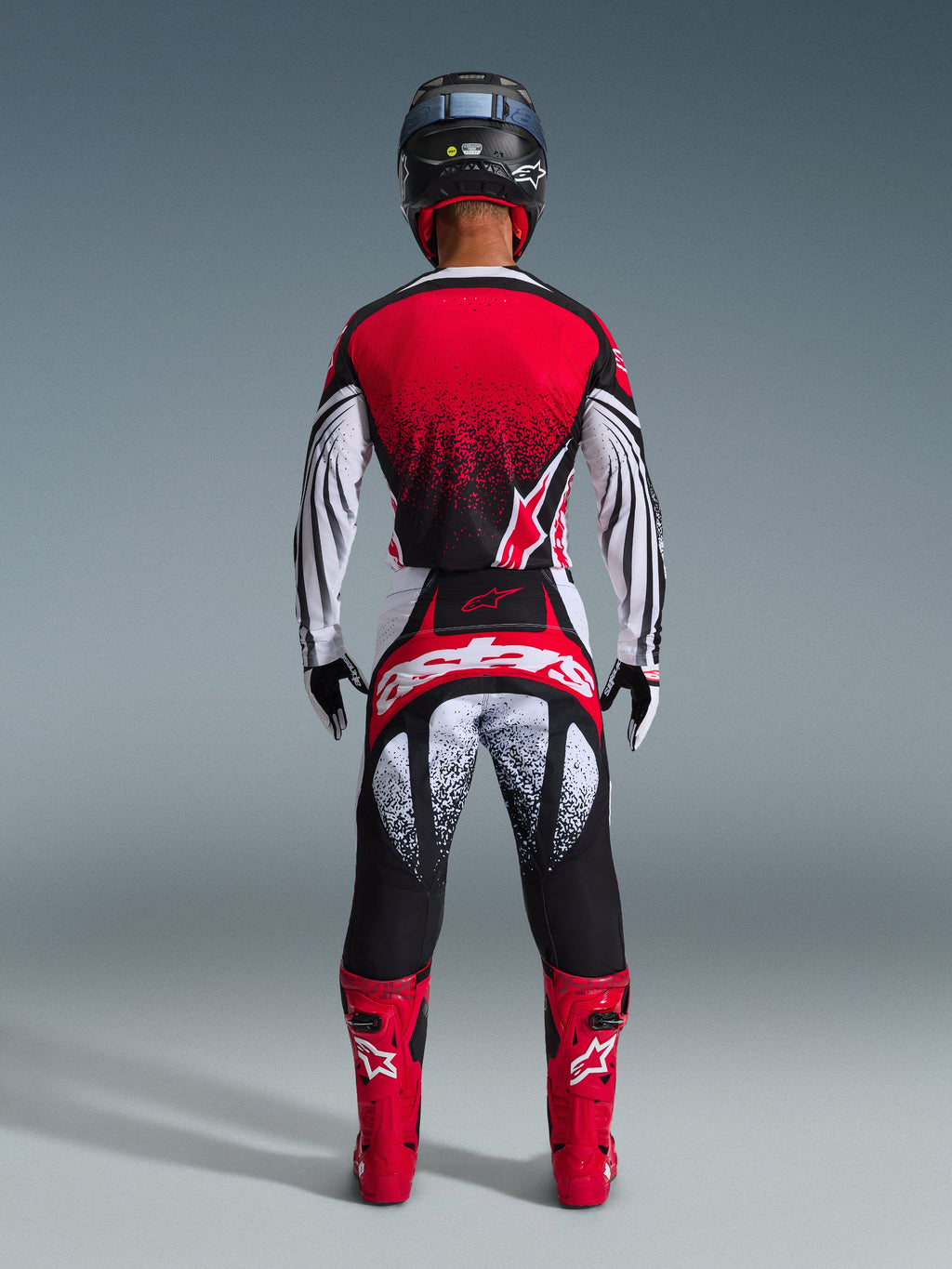 Vista posteriore di un pilota di motocross che indossa i pantaloni Alpinestars Techstar Nomur 2026 in Nero Bianco Rosso, caratterizzati da branding evidente sulla seduta e grafiche puntinate, abbinati a una maglia rossa coordinata e stivali rossi accesi per un kit da gara off-road completo.