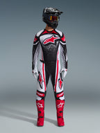 Completo Alpinestars Techstar Nomur 2026, pilota di motocross in piedi con equipaggiamento da gara completo, design nero, bianco e rosso con motivo a puntini bianchi, maglia con logo Astars rosso, pantaloni bianchi e rossi coordinati, stivali rosso acceso e casco nero con maschera, kit da motocross coordinato ad alte prestazioni