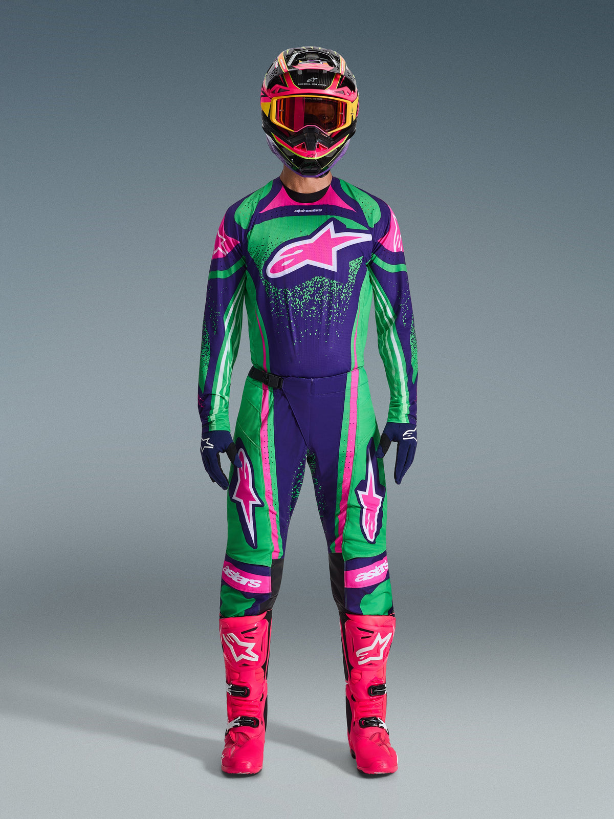 Alpinestars Set Equipaggiamento Techstar Nomur 2026, completo da motocross, verde vibrante, viola e rosa, grande logo a stella su petto e cosce, casco nero con dettagli rosa e gialli, stivali rosa brillante, design sportivo professionale