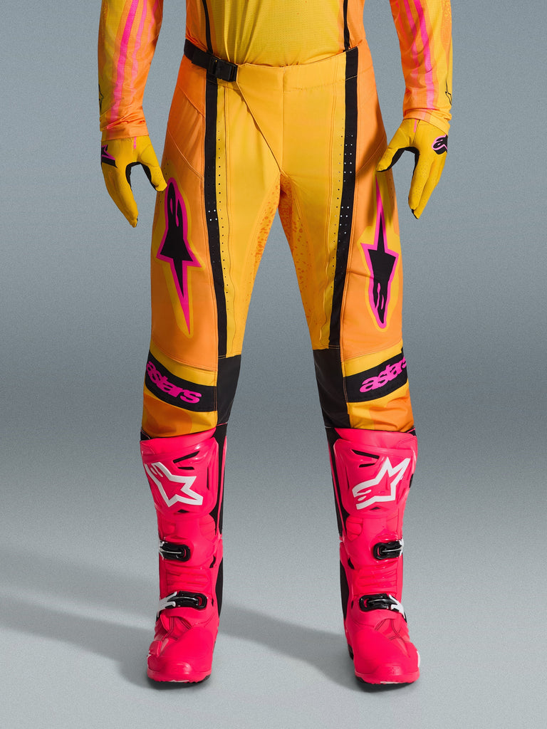 Alpinestars 2026 Techstar Nomur Pants, pantaloni da motocross, arancione, giallo e rosa, indossati con guanti gialli coordinati e stivali da gara rosa acceso, con pannelli neri e vistoso logo Alpinestars rosa, abbigliamento tecnico per competizioni off-road