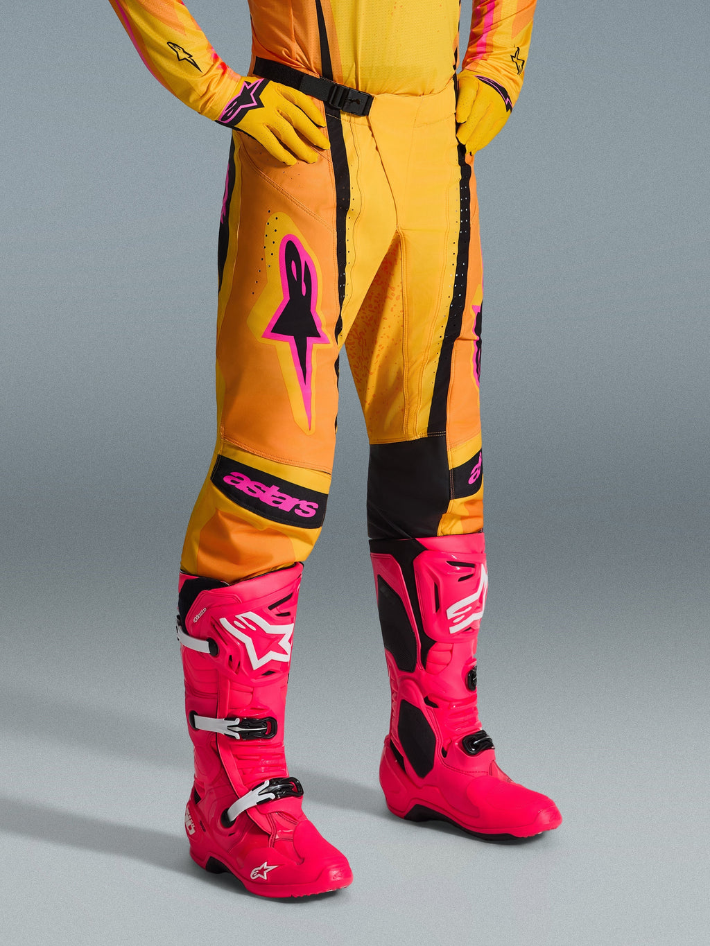 Alpinestars 2026 Techstar Nomur Pants, pantaloni da motocross, arancione, giallo e rosa, indossati con maglia e guanti gialli coordinati e stivali rosa acceso, vestibilità atletica con loghi rosa e fibbia magnetica nera, abbigliamento vivace per competizioni off-road