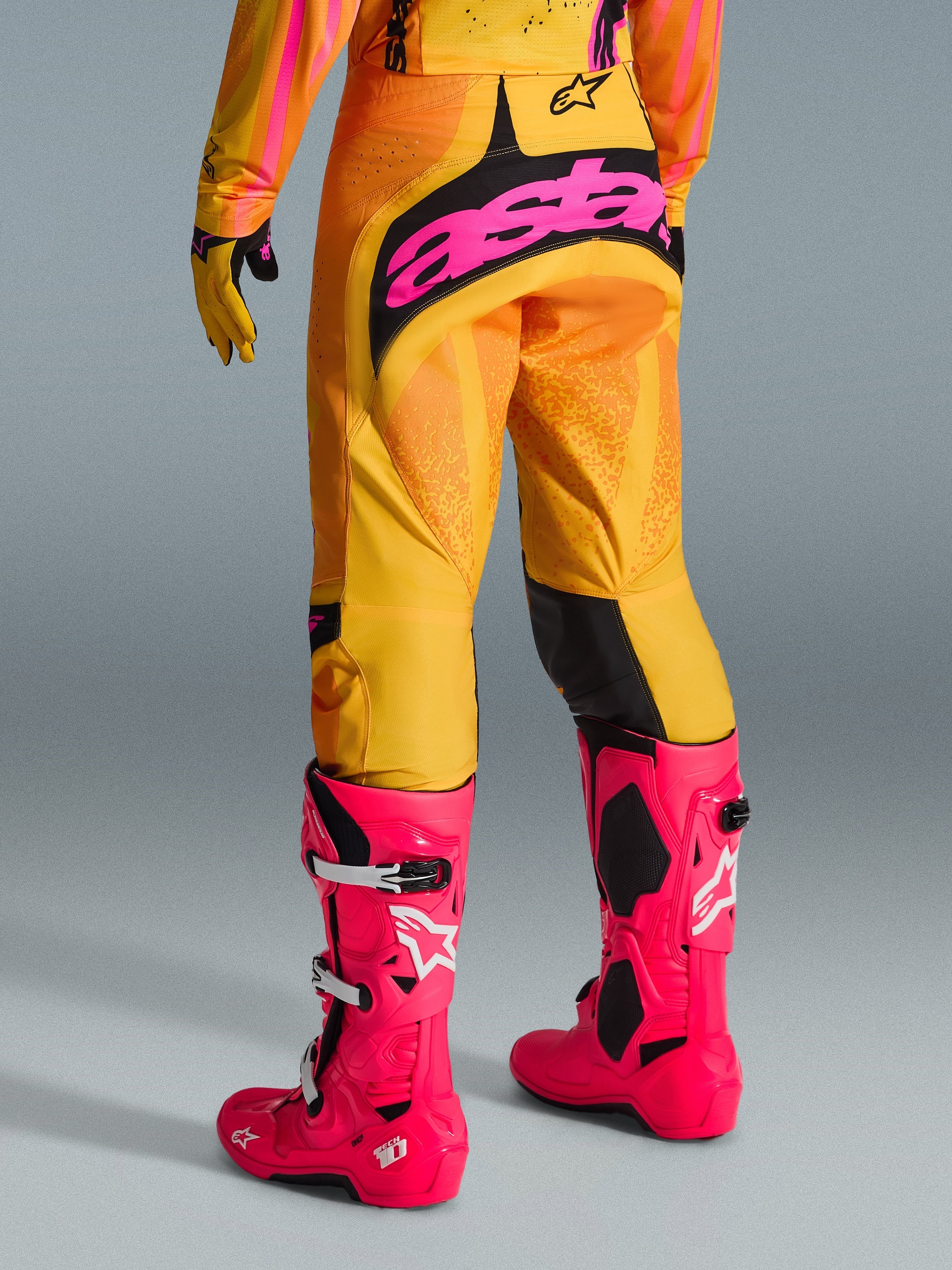 Alpinestars pantaloni Techstar Nomur 2026, pantaloni da motocross, arancione giallo e rosa, vista posteriore con grande logo 'astars' rosa sulla seduta, abbinati a stivali Tech 10 rosa acceso e maglia coordinata, abbigliamento tecnico da gara per motocross