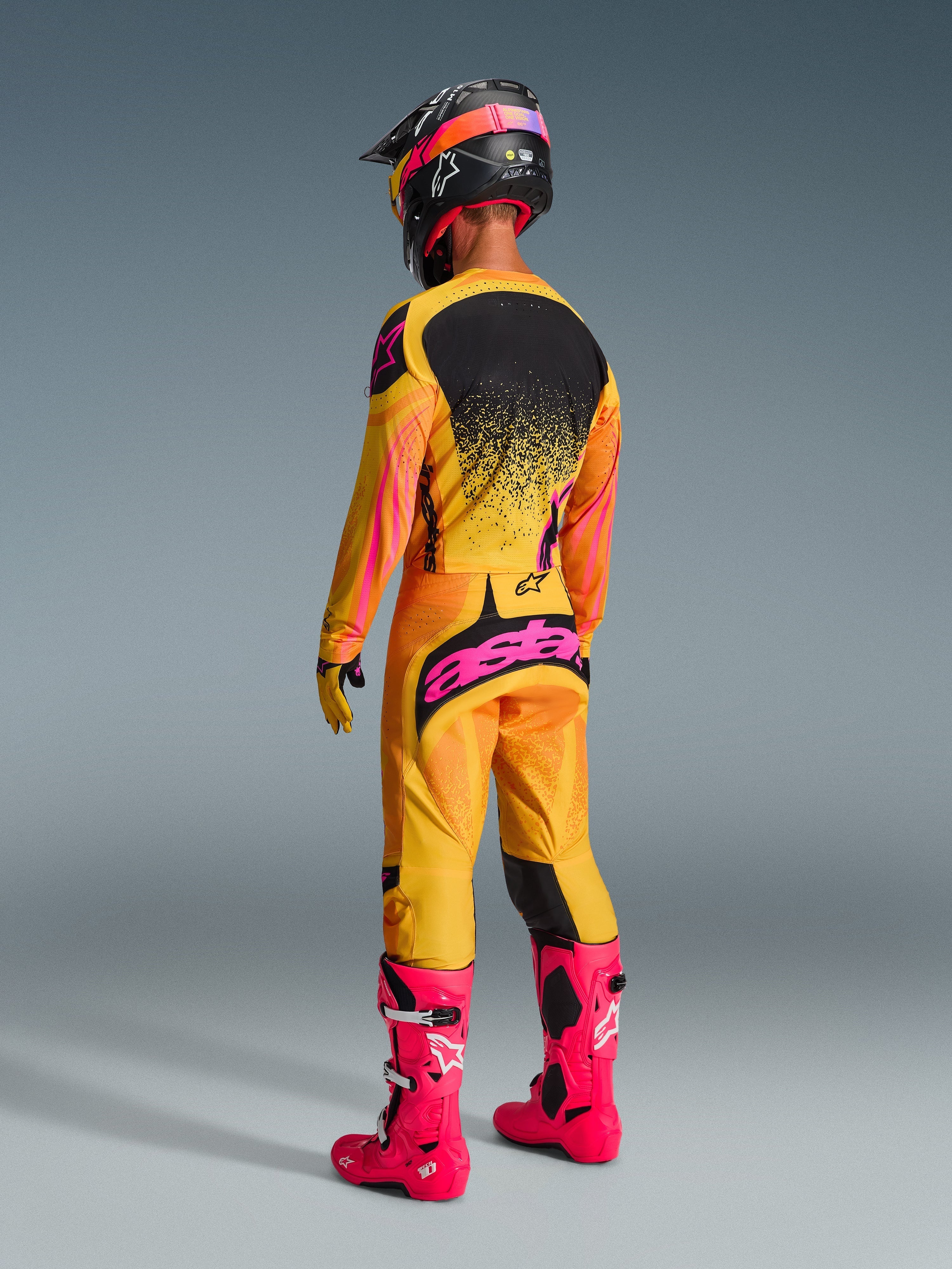 Pilota di motocross visto da dietro con equipaggiamento completo da gara, Pantaloni Techstar Nomur 2026 in arancione, giallo e rosa, vistoso logo 'astars' rosa sulla seduta, maglia coordinata, stivali rosa acceso e casco nero con accenti rosa, completo coordinato per competizioni off-road.