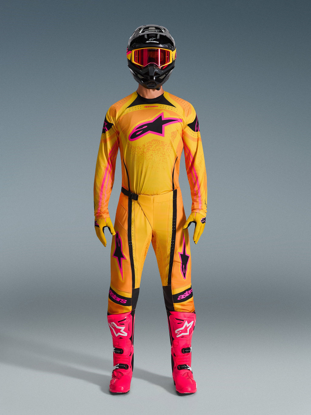 Pilota di motocross con set completo Alpinestars Techstar Nomur 2026, maglia e pantaloni arancioni, gialli e rosa, guanti coordinati gialli e rosa, stivali da corsa rosa acceso e casco nero con occhiali rosa, abbigliamento da motocross coordinato ad alte prestazioni.
