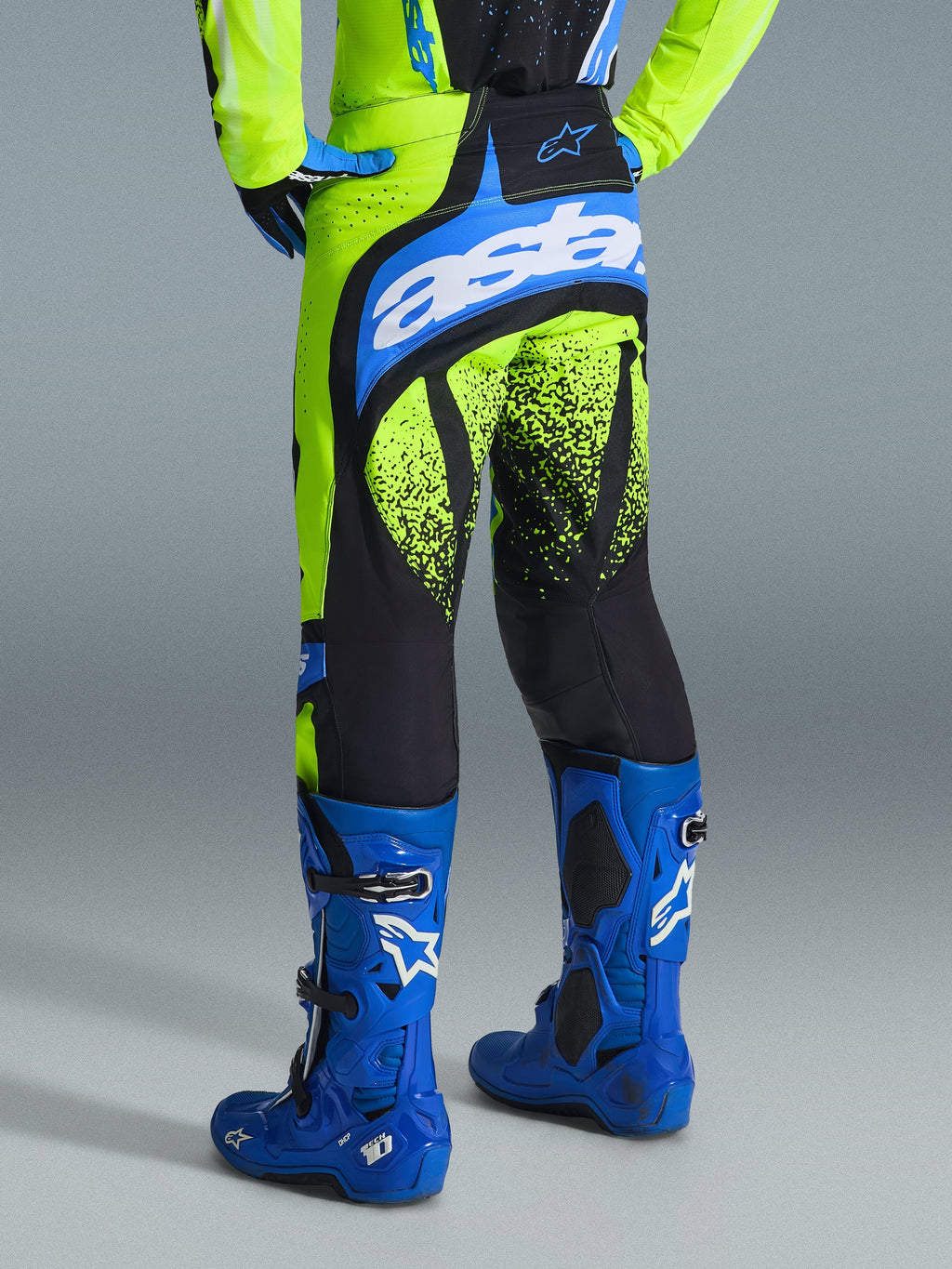 Alpinestars Pantaloni Techstar Nomur 2026, pantaloni da motocross, giallo fluo e blu UCLA, vista posteriore del pilota con stivali blu, caratterizzati dal marchio 'astars' sulla seduta e grafiche nere effetto spruzzo sulle gambe