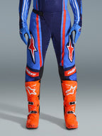 Alpinestars Pantaloni Techstar Nomur 2026, pantaloni da motocross, blu scuro, arancione e blu UCLA, con vistose strisce arancioni e grandi loghi a stella bianchi sulle cosce, indossati con stivali da gara arancione acceso e guanti blu, vestibilità atletica per prestazioni fuoristrada