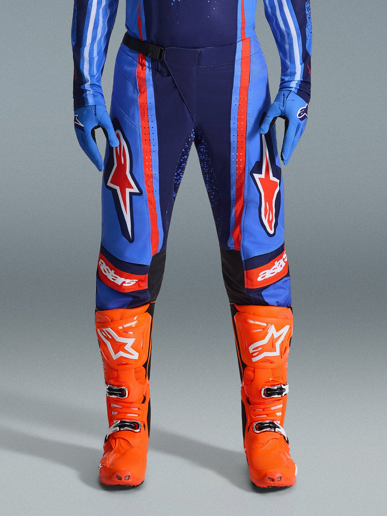 Alpinestars Pantaloni Techstar Nomur 2026, pantaloni da motocross, blu scuro, arancione e blu UCLA, con vistose strisce arancioni e grandi loghi a stella bianchi sulle cosce, indossati con stivali da gara arancione acceso e guanti blu, vestibilità atletica per prestazioni fuoristrada