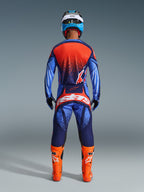 Pilota di motocross visto di spalle con pantaloni Alpinestars Techstar Nomur 2026 in Dark Navy, arancione e UCLA Blue, abbinati a maglia coordinata e stivali arancione acceso, set di abbigliamento professionale da gara off-road.