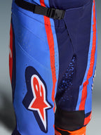 Alpinestars Techstar Nomur Pants 2026, pantaloni da motocross in blu navy scuro, arancione e blu UCLA, primo piano che mostra la chiusura in vita con fibbia magnetica SX2, perforazioni laser sulla coscia e l'iconico logo a stella, equipaggiamento da fuoristrada resistente.